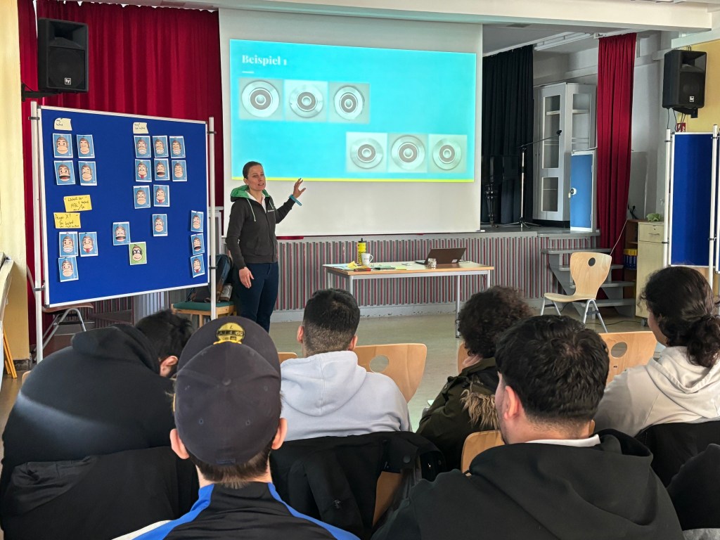 Aus der hinteren Reihe Aufnahme einer Schulklasse. Im unteren Drittel des Fotos sind Hinterköpfe von Schülerinnen und Schülern zu sehen. Vorne vor der Klasse steht Referentin. Links von Referentin ist Pinnwand mit ausgedruckten, vereinfachten Darstellungen von Affenköpfen. Auf dem Beamerbild hinter der Referentin sind sechs Spritzguss-Werkstücke zu sehen.