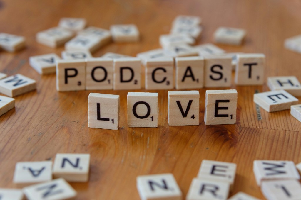 Scrabble-Buchstaben liegen auf Tisch. PODCAST LOVE ist aus einzelnen Buchstaben aufgestellt worden zwischen den anderen liegenden Buchstaben